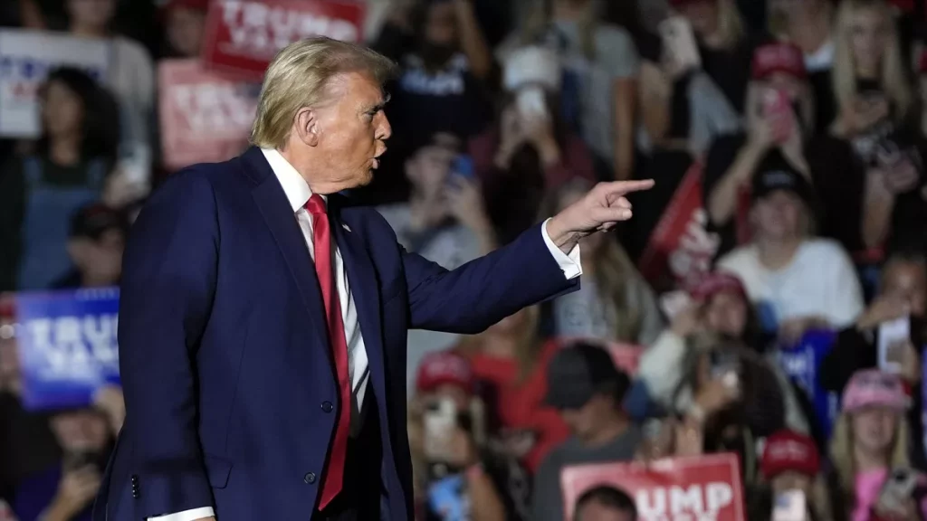 Un homme en costume parle avec enthousiasme à une foule, pointant du doigt, entouré de personnes brandissant des pancartes et portant des chapeaux rouges.