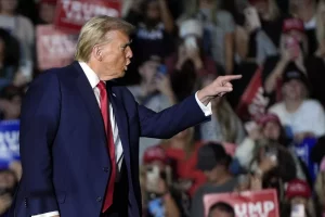 Un homme en costume parle avec enthousiasme à une foule, pointant du doigt, entouré de personnes brandissant des pancartes et portant des chapeaux rouges.