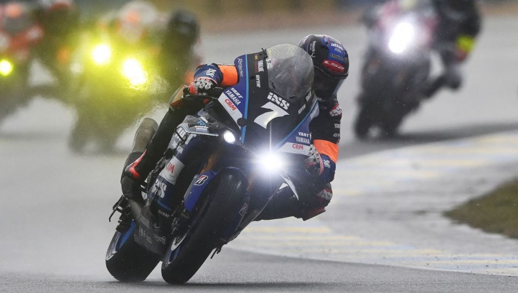 Un pilote en pleine compétition sur sa moto, avec d'autres coureurs en arrière-plan, tous roulant sous la pluie.