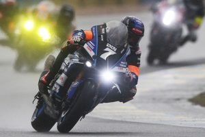Un pilote en pleine compétition sur sa moto, avec d'autres coureurs en arrière-plan, tous roulant sous la pluie.