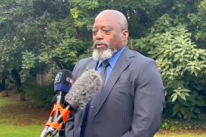 Homme parlant devant des caméras dans un jardin, vêtu d'un costume noir et cravate bleue.