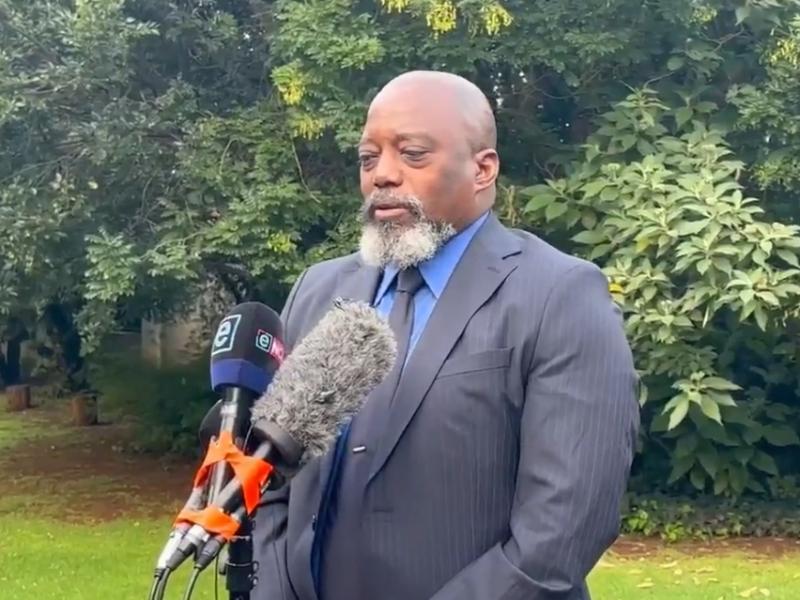 Homme parlant devant des caméras dans un jardin, vêtu d'un costume noir et cravate bleue.