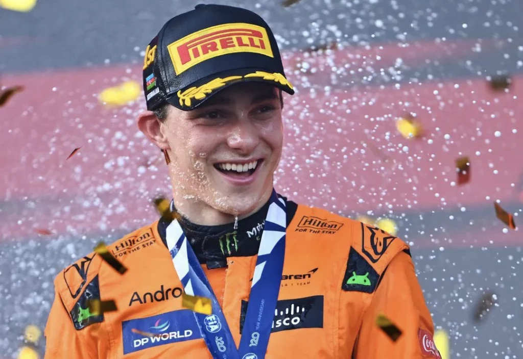 Un jeune pilote souriant en combinaison orange, célébrant sa victoire avec des confettis et de l'eau, portant une casquette Pirelli et une médaille autour du cou.