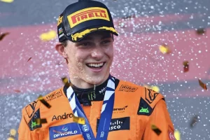 Un jeune pilote souriant en combinaison orange, célébrant sa victoire avec des confettis et de l'eau, portant une casquette Pirelli et une médaille autour du cou.