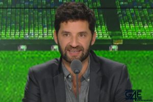 Un animateur de télévision souriant, vêtu d'une veste grise, parlant à un micro devant un fond vert flou d'un stade.