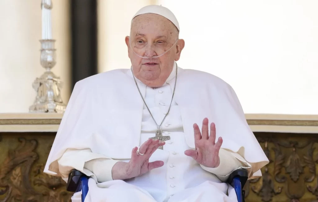 Le Pape François assis, levant les mains pour saluer, avec une attention particulière sur son équipement médical et sa tenue blanche.