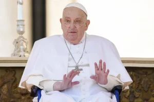 Le Pape François assis, levant les mains pour saluer, avec une attention particulière sur son équipement médical et sa tenue blanche.