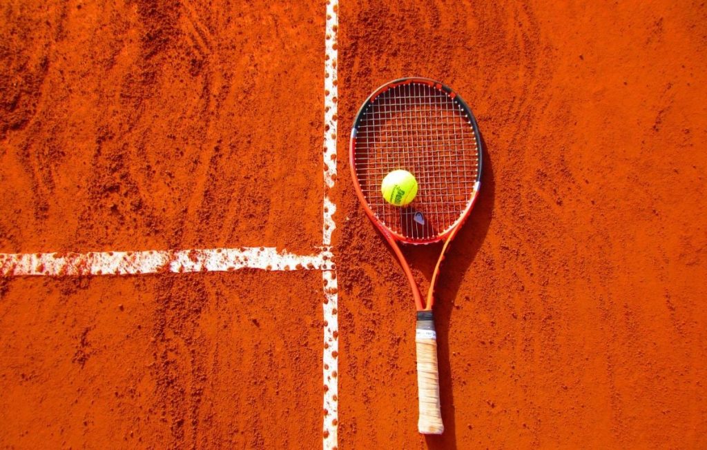 Une raquette de tennis orange et une balle jaune reposent sur un court de tennis en terre battue rouge, avec des lignes blanches visibles.