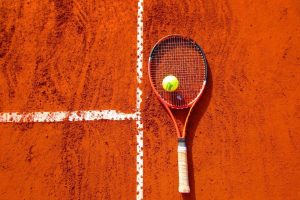 Une raquette de tennis orange et une balle jaune reposent sur un court de tennis en terre battue rouge, avec des lignes blanches visibles.