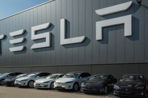 A line of Tesla cars parked in front of a large Tesla logo on a modern building.