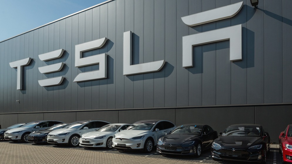A line of Tesla cars parked in front of a large Tesla logo on a modern building.