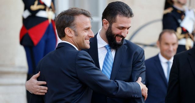 Deux hommes se serrent dans une accolade amicale lors d'un événement officiel, avec des membres des forces de l'ordre en arrière-plan.