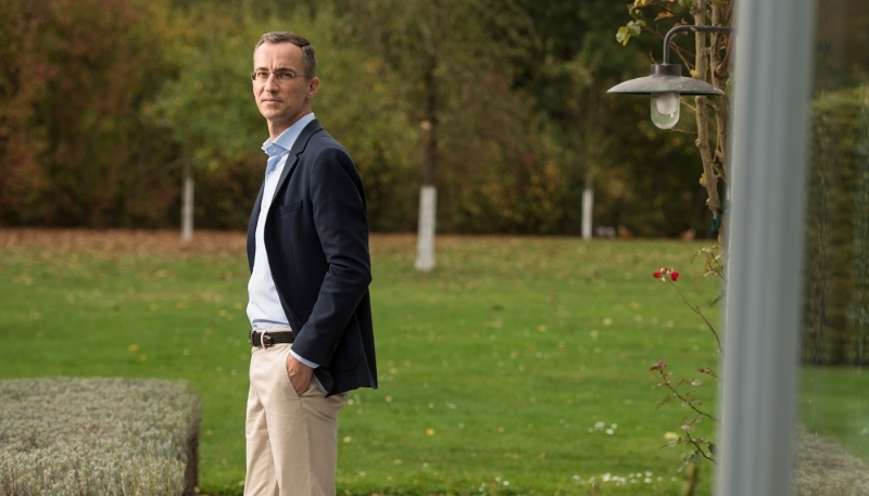 Un homme en costume noir et chemise claire se tient debout dans un jardin avec une expression réfléchie.