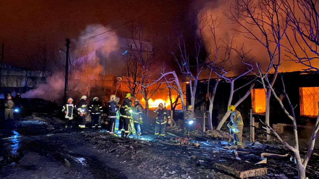 Des pompiers combattent un incendie dans un bâtiment en feu, avec des flammes visibles et de la fumée dans une nuit sombre.