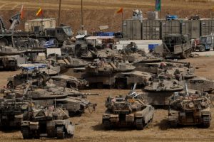 Vue aérienne de plusieurs chars de combat stationnés dans un camp militaire, avec des véhicules de soutien et des conteneurs en toile de fond.