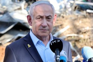 Un homme en costume sombre et chemise bleu clair se tient devant des décombres, exprimant son sérieux lors d'une déclaration publique.